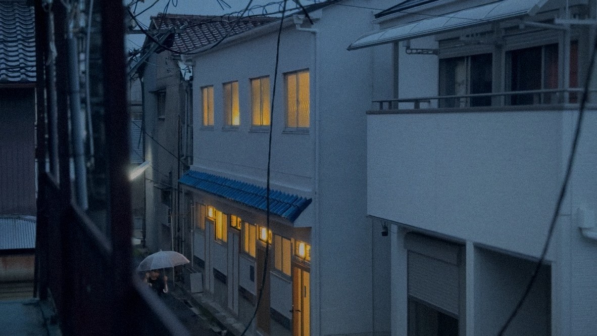 HERE Senbonkita at dusk, its windows lit under the blue tiled awning, seen from across a narrow lane in the rain