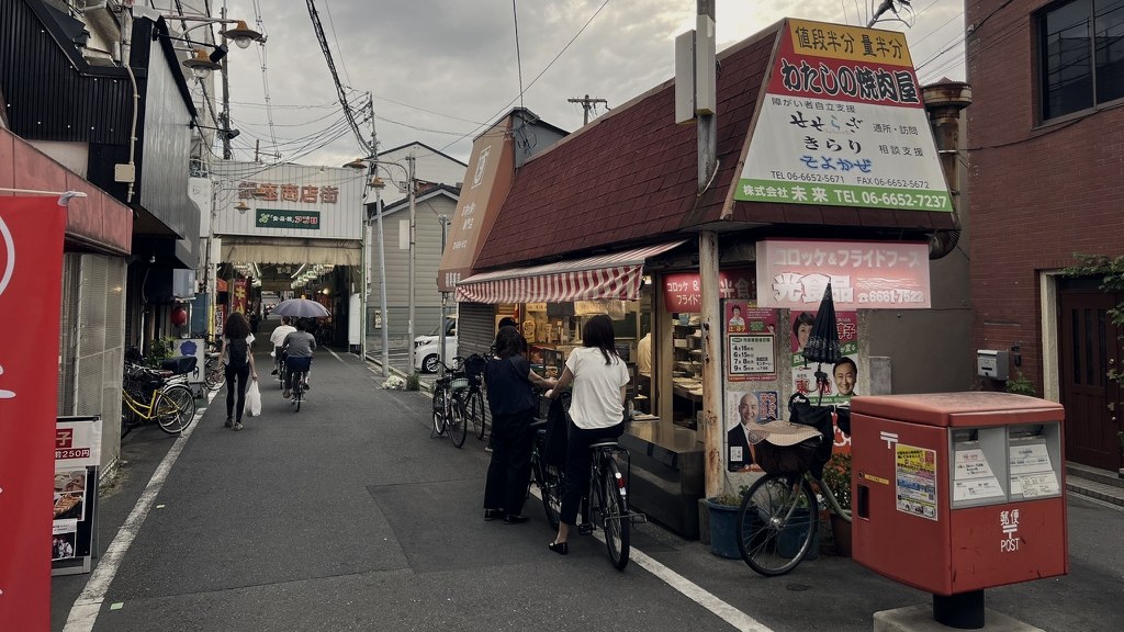 A street corner at dusk: a croquette shop, a red post box and people on bicycles
